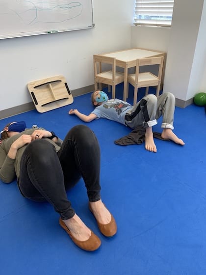 woman laying on blue mat with young boy doing therapeutic activtities